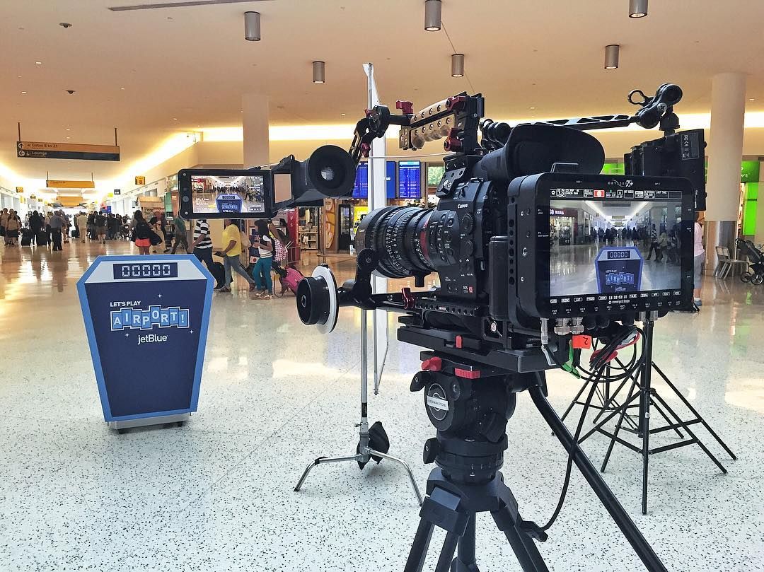 C500 on tripod in the JFK terminal with the "Let's Play Airport!" jetBlue podium in frame, monitors showing the shot